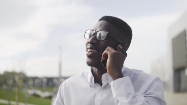 Joven empresario africano con aparatos ortopédicos hablando por teléfono celular en el parque de la ciudad — Vídeos de Stock