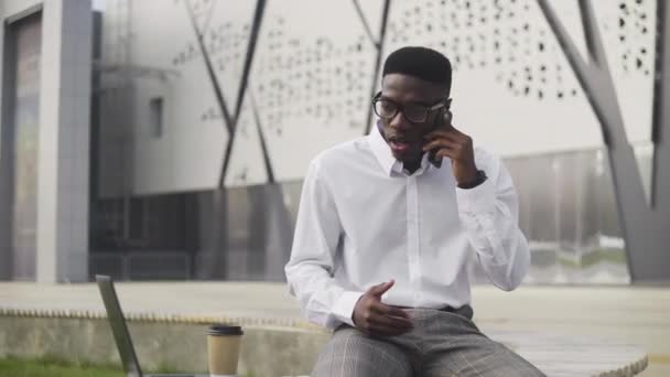 Poważny afrykański biznesmen w okularach rozmawiający przez telefon komórkowy w parku miejskim — Wideo stockowe