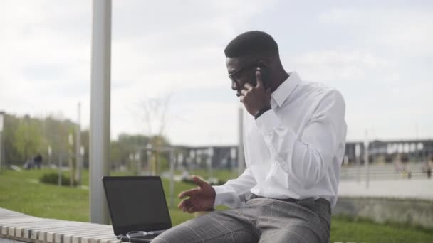 도시 공원에서 휴대 전화로 이야기하고 커피를 마시는 안경을 가진 아프리카 사업가의 측면보기 — 비디오