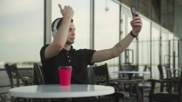 Joven con auriculares en la cabeza haciendo selfie utiliza teléfono sentado en la cafetería — Vídeo de stock