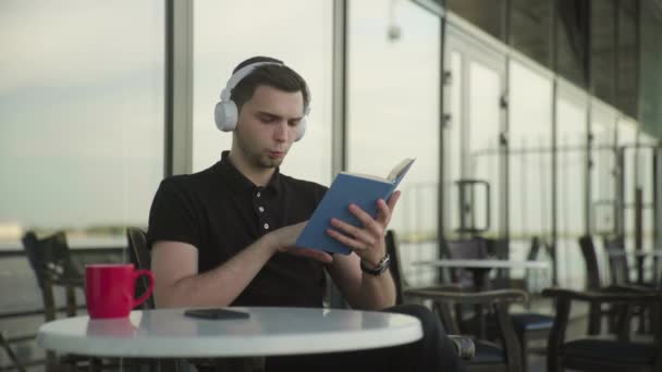 Nuori komea mies istuu puistossa kahvilassa lukemassa kirjaa — kuvapankkivideo