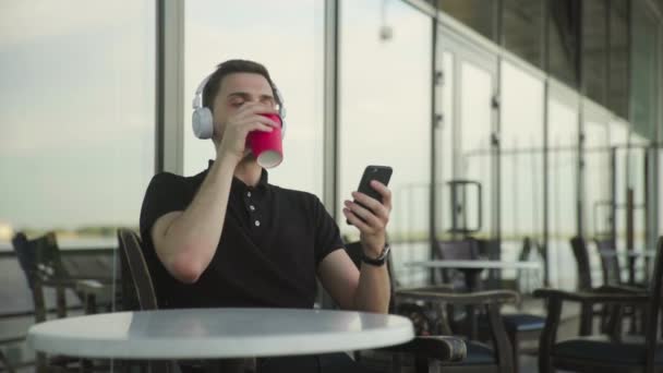 Jóképű fiatalember fülhallgatóval telefonnal ül a kávézóban. — Stock videók