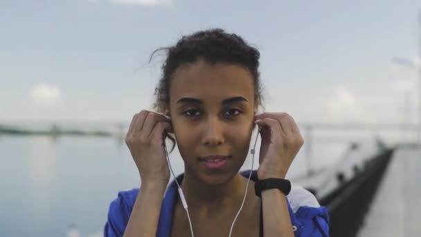 Mujer afroamericana joven está mirando a la cámara y usando auriculares para la motivación musical para el entrenamiento cardiovascular, río en el fondo — Vídeos de Stock