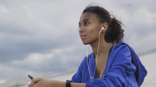 Женщина в спортивной одежде, слушает музыку с мобильного телефона в наушниках, между занятиями в парке — стоковое видео