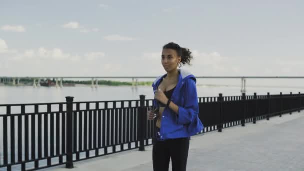 Mujer corredora afroamericana en chaqueta azul con auriculares a lo largo del terraplén — Vídeos de Stock