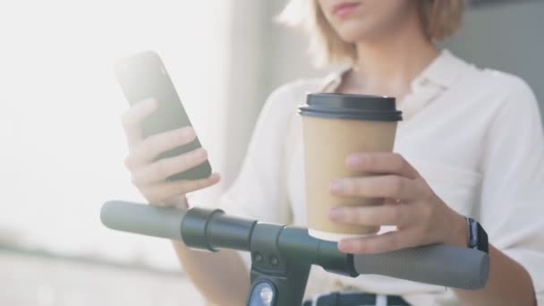 女性は電動キックスクーターに乗ってからスマートフォンやコーヒーを飲んでいます — ストック動画