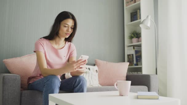 Vonzó fiatal nő gépel az okos telefonján, miközben otthon ül a kanapén.. — Stock videók