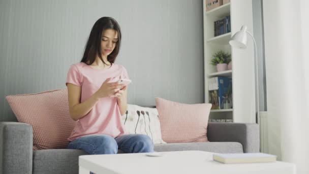Молода сексуальна брюнетка використовує телефон, сидячи на дивані вдома . — стокове відео