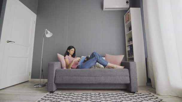 Сонная женщина лежит на диване и читает скучную книгу дома. Студентка колледжа учится на диване глядя учебник чувствую себя скучно — стоковое видео