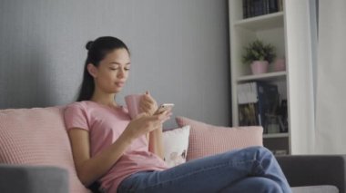 Güzel genç bir kadın akıllı telefon kullanıyor. Kanepede dinleniyor. İnternetten mesaj atıyor, kahve içiyor, rahat bir hayat sürüyor.