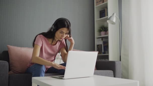 Kulaklıklı genç bir kadın ekran laptopuna bakıyor ve klavyede daktilo yazıyor. — Stok video