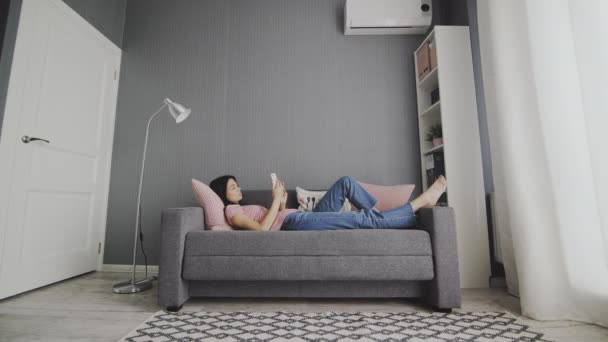 Beautiful young woman holding a smartphone in hands and typing on its screen while chatting in the living room at home. — Stock Video