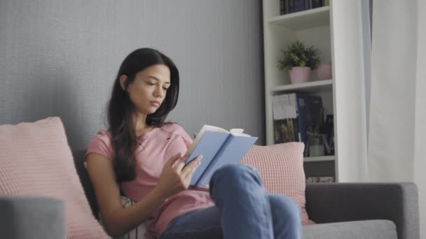 Hermosa joven leyendo un libro triste mientras está sentada en el sofá — Vídeos de Stock