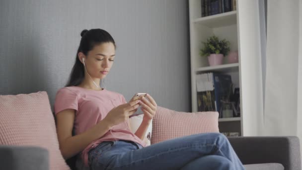Joven mujer morena encantadora sentada en el sofá gris con almohadas rosas, escuchando la música en el teléfono inteligente a través de auriculares blancos — Vídeos de Stock