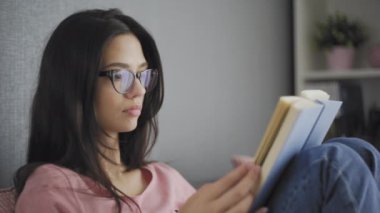 Oldukça güzel, mutlu, gözlüklü bir bayan öğrenci modern bir evin kanepesinde kitap okuyor..