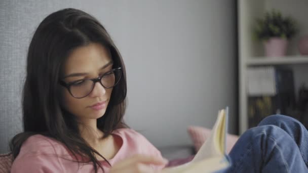 Ziemlich schöne, glückliche Studentin mit Brille, die auf der Couch eines modernen Hauses ein Buch liest. Schauen. Zurück zur Schule. Bildungsuniversität. — Stockvideo