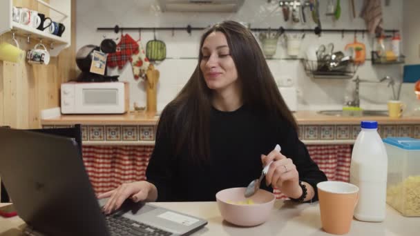 Mujer joven que trabaja ordenador portátil y come cereales de maíz en la cocina casera. — Vídeos de Stock