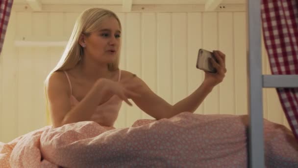 ユースホステルの部屋のベッドに座っている若い魅力的な女性の閉じるとビデオチャットを持っているスマートフォンを使用して — ストック動画