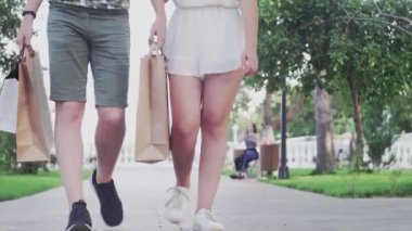 Genç çift ellerinde alışveriş torbalarıyla yaz parkında yürüyor.