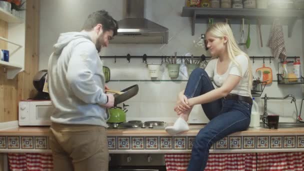 Молодий чоловік готує щось на сковороді, а його дружина сміється з нього — стокове відео