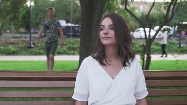 Mladá žena sedí na lavičce a čeká na svého přítele v letním parku — Stock video