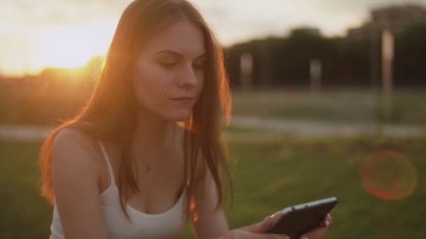 Portret van een jonge vrouw in het park die e-book leest, zonsondergang op de achtergrond — Stockvideo