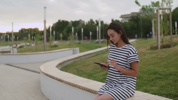 Mujer bastante joven leyendo un libro en el banco al aire libre al atardecer — Vídeo de stock