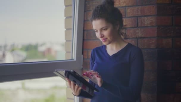 Mooi meisje staat naast het raam en gebruikt de tablet — Stockvideo