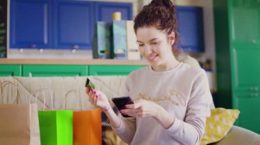 Kadın kanepeye oturmuş alışveriş torbalarıyla internetten alışveriş yapıyor kredi kartı, cep telefonu ve cep telefonu uygulaması kullanıyor.