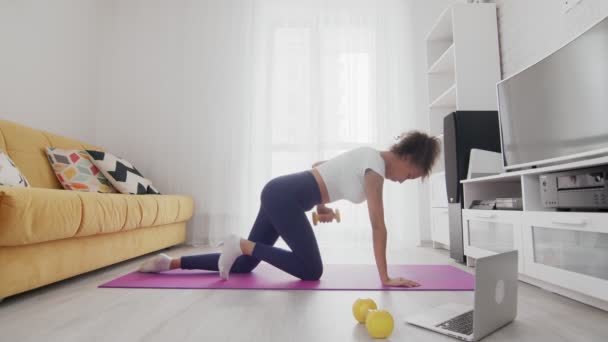 Mladá sportovní africká americká žena vytáhnout žlutý činka na podložce s pomocí svého instruktora on-line na notebooku doma — Stock video