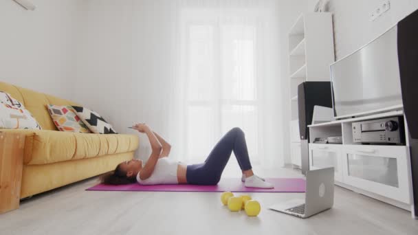 Mladá sportovní africká americká žena leží na podložce a používá telefon po cvičení doma — Stock video