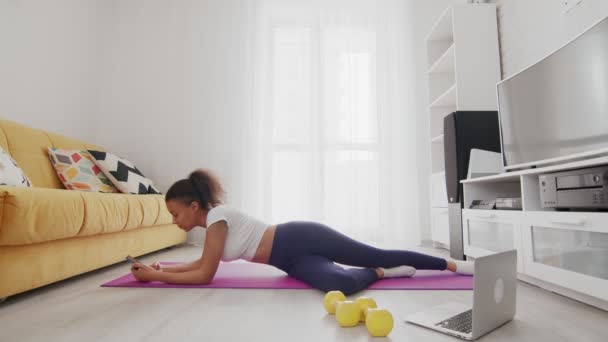 Young sporty african american woman lays down on mat and uses phone after exercises at home — Stock Video