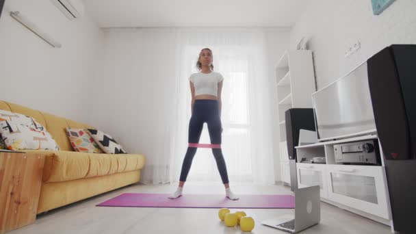 En ung, sportslig, amerikansk kvinne gjør ingenting med et elastisk band på matta ved hjelp av instruktøren på PC hjemme – stockvideo