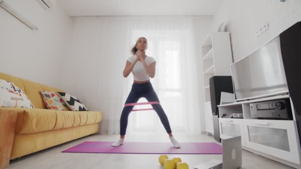 En ung, sportslig, amerikansk kvinne gjør trinn med et elastisk band på matta med hjelp av instruktøren på PC hjemme – stockvideo