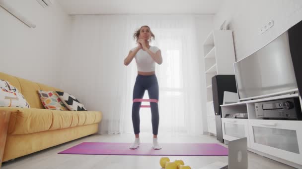 Young sporty african american woman does squats and jumps with elastic band on mat with help of her instructor online on laptop at home — Stock Video