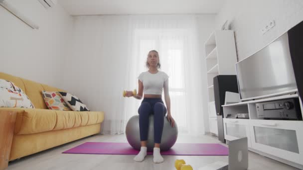 En ung, sportslig, amerikansk kvinne sitter på fitball og trener biceps med dumklokker ved hjelp av instruktøren på PC hjemme – stockvideo