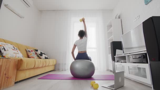 Vista posterior de la joven afroamericana deportiva se sienta en el fitball y tríceps derecho de entrenamiento con pesas con la ayuda de su instructor en línea en el ordenador portátil en casa — Vídeo de stock