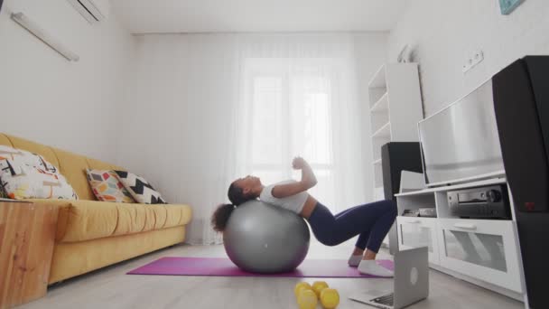 Vista posteriore della giovane donna afroamericana sportiva fa esercizi di fitball con l'aiuto del suo istruttore online sul computer portatile a casa — Video Stock