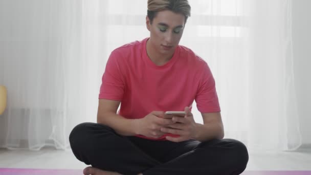 Молодой гомосексуалист с макияжем сидит на коврике Фитнес и начинает пользоваться телефоном — стоковое видео