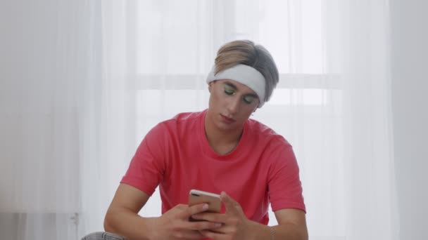 Jeune homme homosexuel transgenre avec maquillage se trouve sur fitnes mat et en utilisant le téléphone — Video