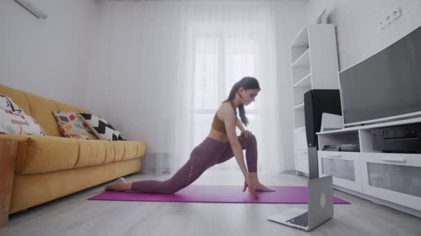 Junge schöne Frau macht Stretching-Übungen auf violetten Matten zu Hause mit Hilfe seines Instruktors online auf Laptop zu Hause — Stockvideo