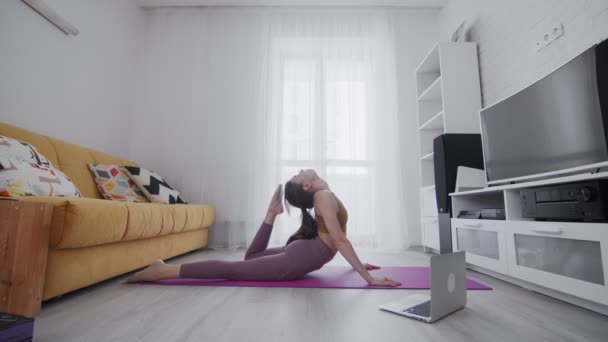Joven hermosa mujer está haciendo ejercicios de estiramiento en estera violeta en casa. Toca la cabeza con el pie — Vídeos de Stock