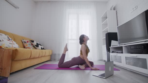 Young beautiful woman is doing stretching exercises on violet mat at home. She touches head with her foot with help of his instructor online on laptop at home — Stock Video