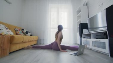 Genç ve güzel bir kadın evde dizüstü bilgisayarında eğitmeninin yardımıyla menekşe minderi üzerinde bacak egzersizleri yapıyor.