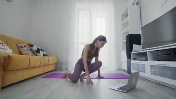 En ung, vakker kvinne trener på en fiolett matte med instruktøren på PC hjemme. – stockvideo