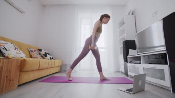 Junge schöne Frau mit Tätowierung macht Dumbbell Lunges auf Matte mit Hilfe ihres Instruktors online auf dem Laptop zu Hause — Stockvideo