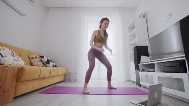Young beautiful woman does squats with help of his instructor online on laptop at home — Stock Video