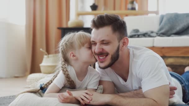 Щасливий батько і його дочка лежать на підлозі. Маленька мила дівчинка цілує свого батька — стокове відео