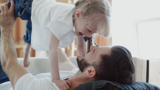 Primer plano del joven en jeans y camiseta blanca juega con su hija en el sofá — Vídeos de Stock