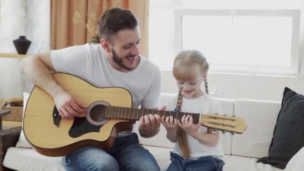 Padre joven enseña a su hija a tocar la guitarra en casa — Vídeos de Stock